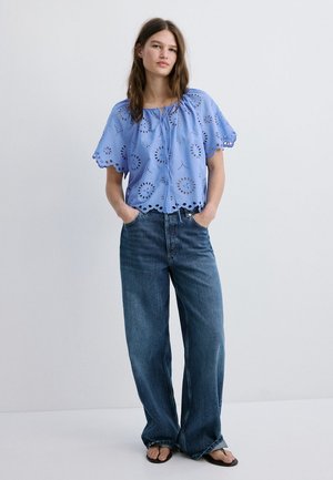 Jeune femme portant un chemisier bleu à œillets et un jean à jambes larges, debout les mains dans les poches devant un fond uni.