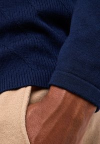 Maglione lavorato a maglia blu navy con motivo testurizzato e polsini a costine, abbinato a un pantalone di stoffa beige chiaro, con una mano appoggiata in una tasca.
