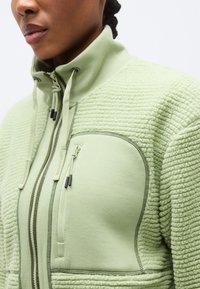 Ανοιχτό πράσινο φλις σακάκι με ψηλό γιακά, κλείσιμο με φερμουάρ και υφή υφάσματος. Διαθέτει τσέπη στο στήθος με φερμουάρ και αντίθετες λεπτομέρειες.