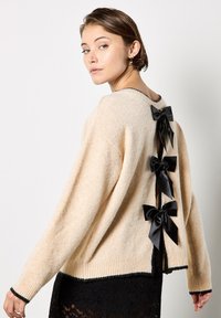 Suéter de punto beige con un corte relajado, que presenta un dobladillo acanalado y tres lazos de satén negros en la parte posterior, resaltando un acabado texturizado.