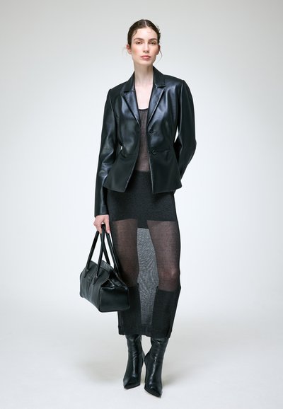 Femme en blazer en cuir noir, jupe noire transparente, bottes noires pointues, tenant un sac à main noir, debout devant un fond blanc uni.