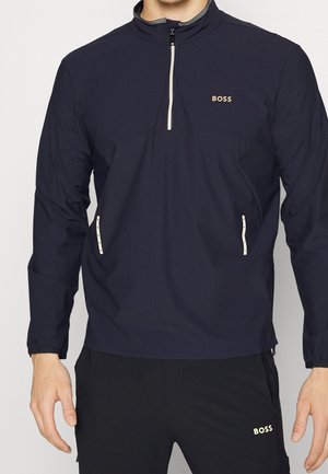 Mann trägt eine marineblaue Quarter-Zip-Jacke und schwarze Hose, beide mit "BOSS" in beiger Schrift.