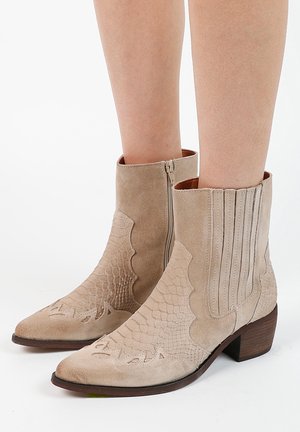 Bottines beiges en daim avec motif serpent texturé, fermeture éclair latérale, petit talon empilé, portées sur jambes nues sur fond blanc.