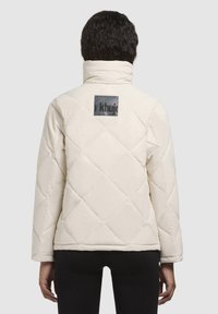 Chaqueta acolchada color crema con un patrón de rombos, cuello alto y un parche gris con el logotipo en la parte superior de la espalda. Tejido texturizado y suave.