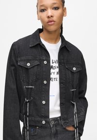 Črna denim jakna s skrajšano zasnovo, sprednjim gumbom, dvema prsnikoma in kontrastnimi belimi logotipnimi detajli blizu roba ter na rokavih.