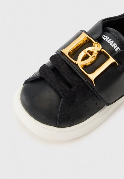 Svart lädersneaker med en vit gummisula, som har en framträdande spänne i guldton och svarta snörning. Strukturerad yta med perforeringar.