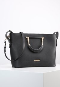 Svart syntetläderhandväska med guldfärgade detaljer, dubbla handtag och avtagbar axelrem. Har en strukturerad form och texturerad yta.