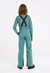 Pantalons de neige teal avec des bretelles noires ajustables, des jambes fuselées et une texture lisse, associés à un haut teal à manches longues. Chaussures noires.