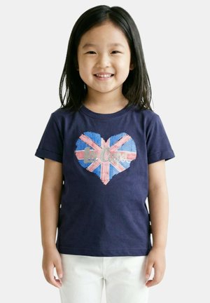 Jeune fille souriante aux cheveux noirs raides portant un t-shirt bleu marine avec un drapeau britannique en forme de cœur et un pantalon blanc, debout devant un fond blanc.