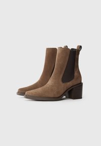 Bottines en daim marron avec un talon carré, dotées de panneaux latéraux élastiques et de languettes pour un enfilage facile. Texture lisse avec des lignes épurées.
