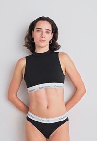 Kvinne med skulderlangt mørkt hår iført svart Calvin Klein ermeløs crop top og matchende svart undertøy med hvite elastiske bånd.