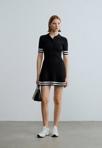 Melna adīta polo kleita ar baltām svītrām uz piedurknēm un apakšmalu. Valkāta ar baltām sporta kurpēm un rokās tur mazu melnu somiņu.