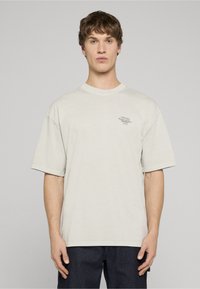 Světle béžové oversized tričko z měkké bavlny s kulatým výstřihem a krátkými rukávy. Na prsou je umístěno malé potištěné logo.