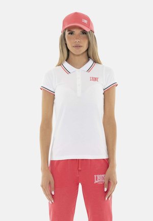 Donna con polo bianca con bordi rossi e neri, cappellino rosso e pantaloni rossi con loghi "Leone 1947", in piedi davanti a uno sfondo neutro.