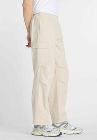 Pantalones cargo beige con cintura elástica, bolsillos laterales y un corte relajado, combinados con zapatillas gris y blancas. Textura suave.