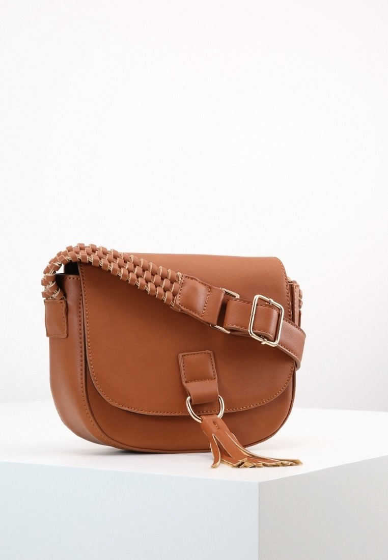 Brun läder crossbody-väska med flätad rem, mässingsdetaljer, klaffstängning och en accenttassel. Slät textur, rundad form.