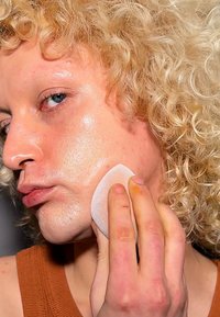 Kihara vaaleat kiharat hiukset, lähikuva kasvoista, joilla on sileä ja kiiltävä iho; käsi pitää pyöreää meikkisovellinta poskea vasten.