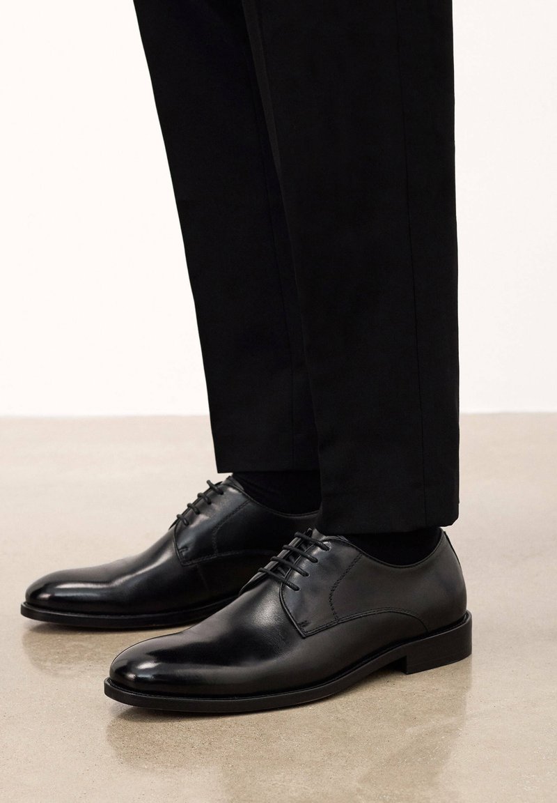 Zapatos de vestir de cuero negro con un acabado liso, punta redonda y seis ojales. Combinados con pantalones de corte negro.