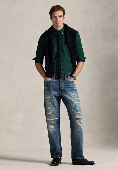 Man wearing green checked shirt with black tie and sweater tied over shoulders, distressed jeans, and black shoes standing with hands in pockets.