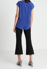 Královsky modré tričko s krátkým rukávem a zaobleným lemem, spárované s černými zkrácenými kalhotami s rozšířením ve spodní části a třásněmi na manžetách, doplněné pruhovanými block-heeled sandály.