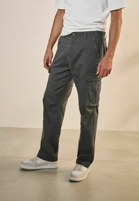 Pantalones cargo grises con bolsillos laterales, diseño de pierna recta, fabricados con tejido duradero, combinados con zapatillas blancas sobre un suelo de concreto claro.