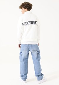 Sudadera blanca con el estampado "LAYERED" en gris en la parte posterior, combinada con jeans cargo de rayón azul claro y zapatillas blancas, vista desde detrás.