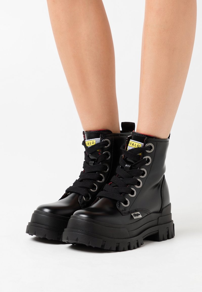 Bottes à plateforme en cuir noir à lacets avec semelles épaisses crantées, portées sur des jambes nues sur fond blanc.