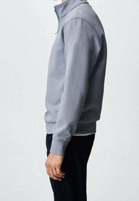 Homme portant un pull-over gris clair à fermeture éclair quart et un pantalon sombre, se tenant de profil avec le bras droit détendu le long du corps.