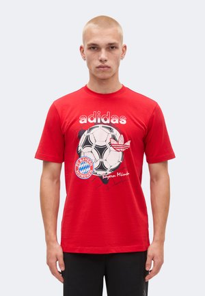Jeune homme portant un T-shirt rouge Adidas avec un ballon de football, le logo du Bayern Munich et un texte sur le devant, debout devant un fond uni.