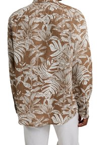 Chemise marron à manches longues avec motif blanc de feuilles et de plantes tropicales portée avec un pantalon blanc, vue de dos d'une personne.