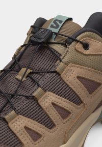 Κοντινή λήψη καφέ και ταμπά ορειβατικού παπουτσιού με αεριζόμενο πλέγμα, σουέντ επικαλύψεις και σύστημα ταχείας απελευθέρωσης κορδονιών σε μαύρο χρώμα στη γλώσσα.