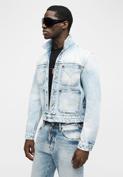 Άνδρας που φοράει ανοιχτό μπλε τζιν μπουφάν με λευκή έκπλυση και τζιν παντελόνι, μαύρο πουκάμισο από μέσα και μεγάλα μαύρα γυαλιά ηλίου, όρθιος σε πλάγια στάση.