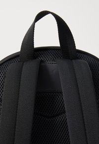 Sac à dos noir avec des bretelles en mesh rembourrées et une poignée sur le dessus pour le transport. Texture lisse avec un tissu durable et des détails texturés.