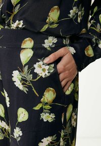 Czarna tkanina z nadrukiem zielonych awokado i białych kwiatów. Ręka spoczywa na bocznej kieszeni. Widoczny detal guzikowego mankietu. Gładka tekstura.