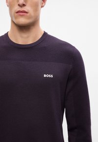 Dunkellila Strickpullover mit gerippter Textur, Rundhalsausschnitt und einem weißen "BOSS"-Logo auf der Brust. Verfügt über klare Linien und eine taillierte Passform.