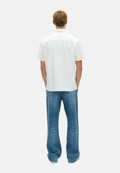 Wit overhemd met korte mouwen en een relaxte pasvorm, gecombineerd met blauwe denim jeans met een wijde pijp. De outfit heeft een simpele, casual uitstraling.