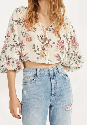 Blusa blanca con estampados florales en rosa y verde, con escote de lazo y mangas abullonadas. Combinada con vaqueros desgastados azul claro.