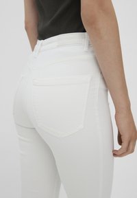 Bílé džíny s vysokým pasem a slim fit střihem, vybavené zadními kapsami a hladkým, elastickým materiálem. Na pasu je přišitá nálepka.
