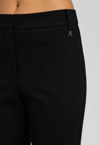 Pantaloni neri su misura realizzati in tessuto liscio, con un discreto logo A in metallo, design slim fit e fronte piatto senza tasche visibili.