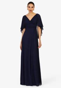 Frau in einem marineblauen, bodenlangen Kleid mit plissiertem V-Ausschnitt und transparenten Umhängeärmeln, stehend vor einem weißen Hintergrund.