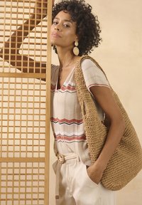 Geweven rieten handtas in beige, grote hobo stijl met een gestructureerde oppervlakte. Model draagt een gestreepte crème blouse en lichte broek met een riem.