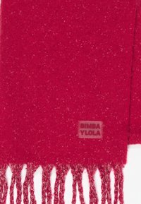 Bufanda de lana roja con extremos flecos y una pequeña etiqueta cosida que dice "BIMBA Y LOLA" cerca del borde.