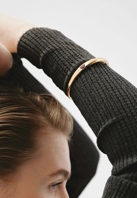 Pulsera de oro con un pequeño gema roja, llevada sobre un suéter negro texturizado. El diseño es suave y redondeado.
