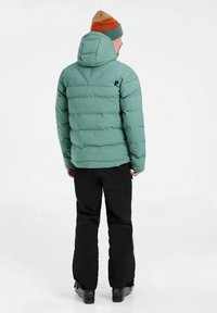 Chaqueta de plumas verde menta con capucha, hecha de material acolchado. Pantalones de esquí negros y botas oscuras completan el atuendo. Gorra de contraste en marrón y verde.