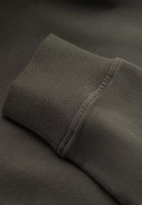 Campione di tessuto verde scuro con polsino a costine. Il materiale è liscio con una leggera lucentezza, evidenziando cuciture piatte e una trama fine.