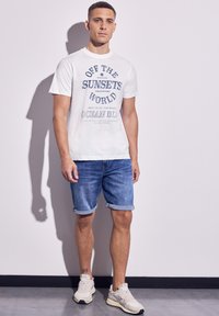 Vit grafisk t-shirt med blå text, tillsammans med upprullade jeansshorts och ljusa sneakers. Skuggor skapar djup mot en enkel bakgrund.