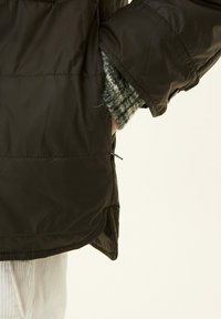 Dark insulated jacket with a smooth, shiny finish. Features a high collar and side pocket, layered over a textured green knit sweater.