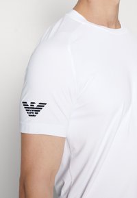 Vit idrottströja med korta ärmar, med en texturerad tyg och en svart präglad logotyp på högra ärmen.