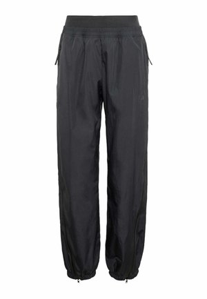 Personne souriante portant une veste polaire noire à capuche, un pantalon noir, des chaussures noires et un sac à dos noir, debout devant un fond uni.