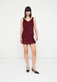 Moteris, vilkinti be rankovių, tekstūruotą tamsiai raudoną mini suknelę, laiko šviesiai rožinę delninę, vilkinti juodais slingback aukštakulniais, stovinti ant baltos grindų.
